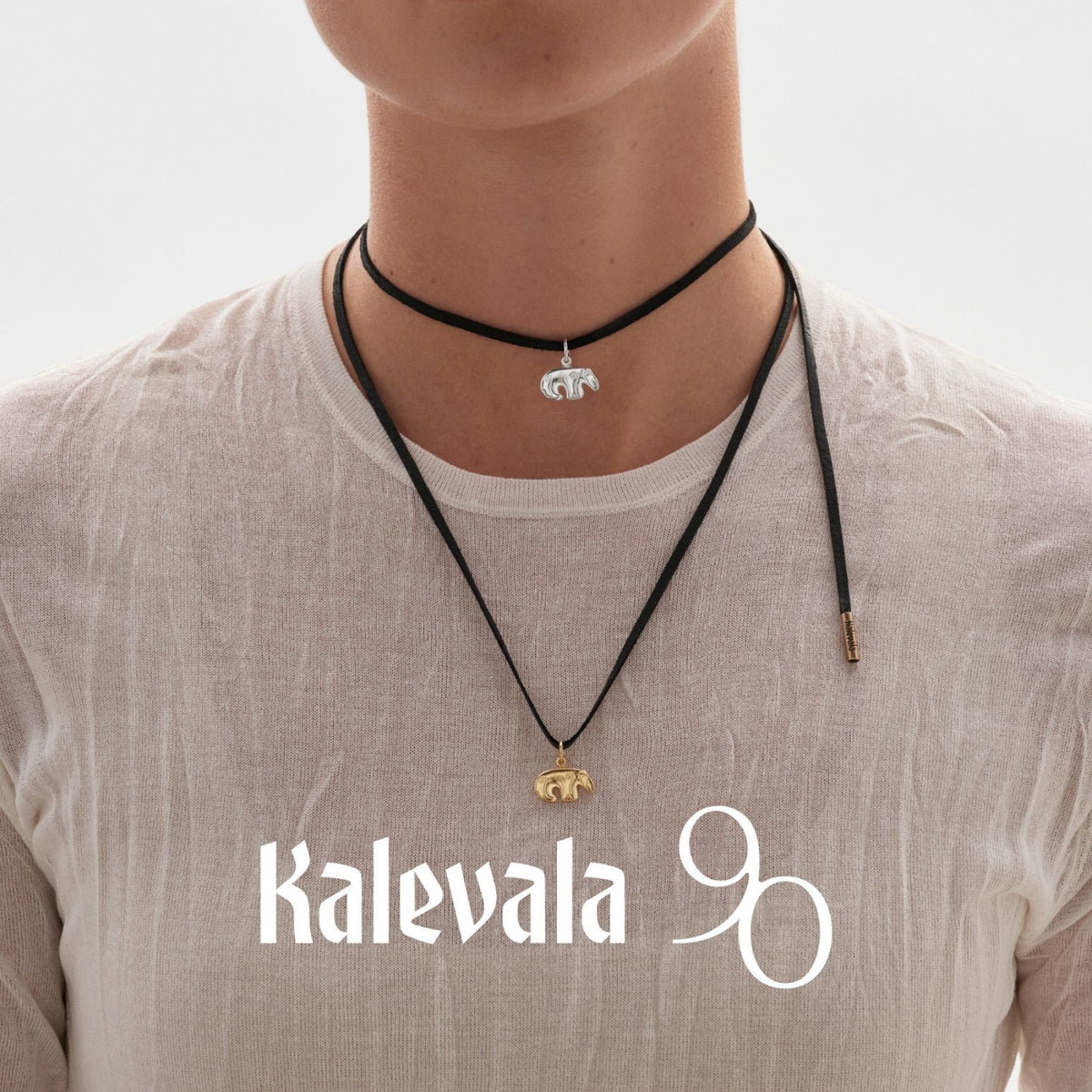 Kalevalan lämminsävyinen pronssinen karhuhela mustassa kaulanauhassa, mallina valkoiseen puseroon pukeutunut henkilö ja Kalevala 90 -tunnus