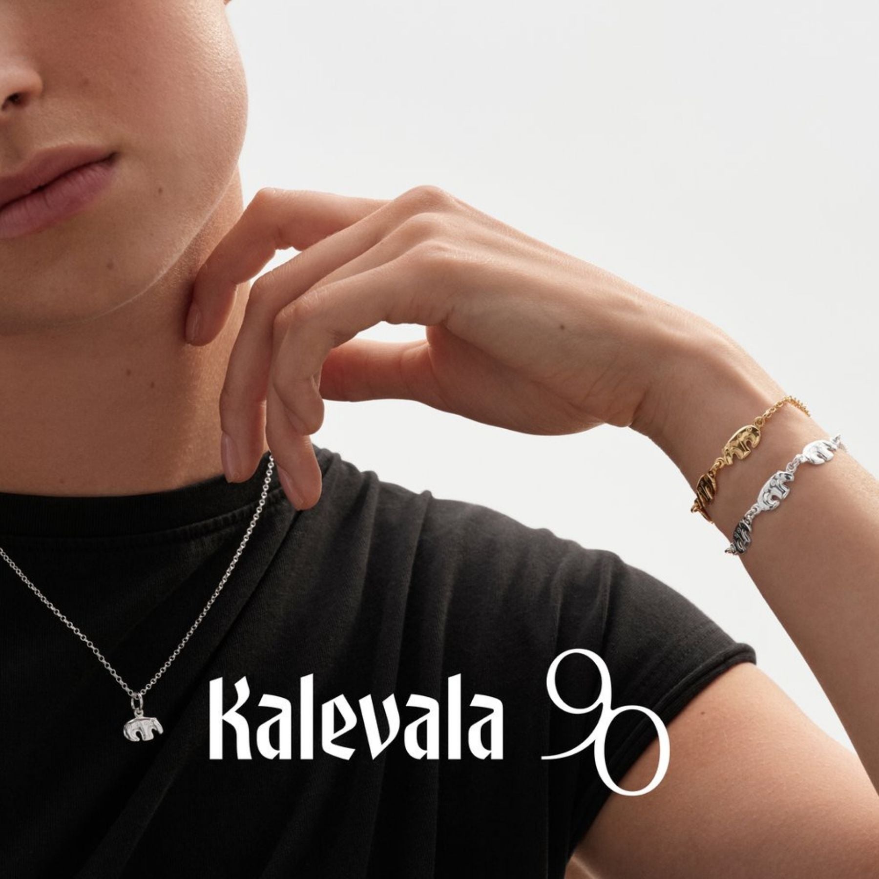 Kalevalan hopeinen karhuriipus ketjussa mustaa paitaa vasten, ranteella kaksi rannekorua ja Kalevala 90 -tunnus