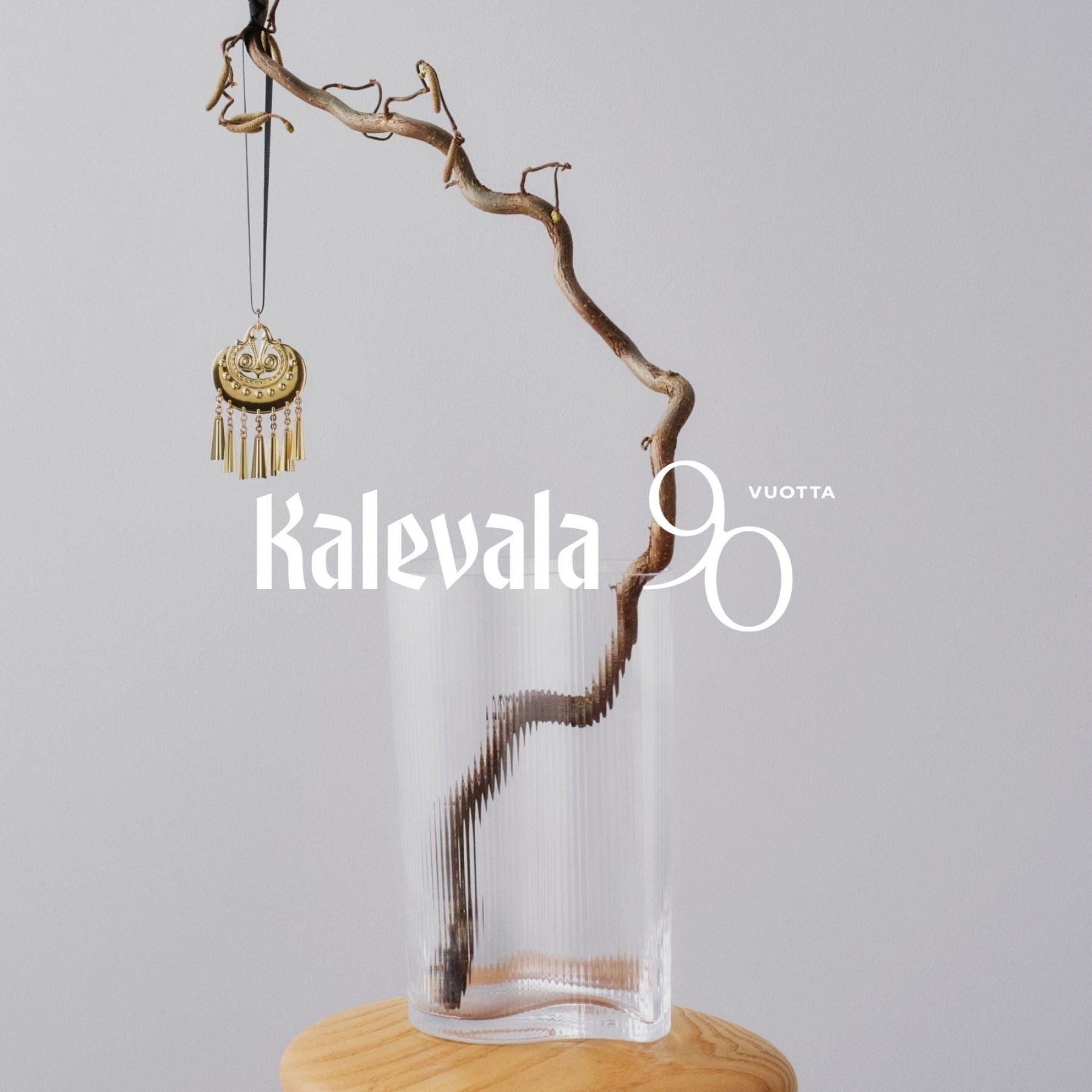 Kalevalan kullanhohtoinen pronssikoriste roikkuu mustasta nauhasta oksalla lasimaljakon yllä.
