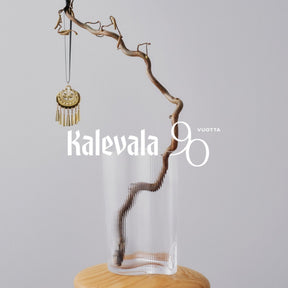 Kalevalan kullanhohtoinen pronssikoriste roikkuu mustasta nauhasta oksalla lasimaljakon yllä.